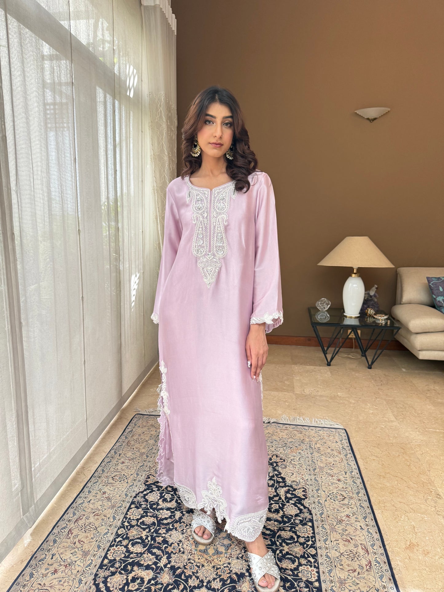 Woman in a light purple dress standing in a room with a beige wall and a patterned rug.