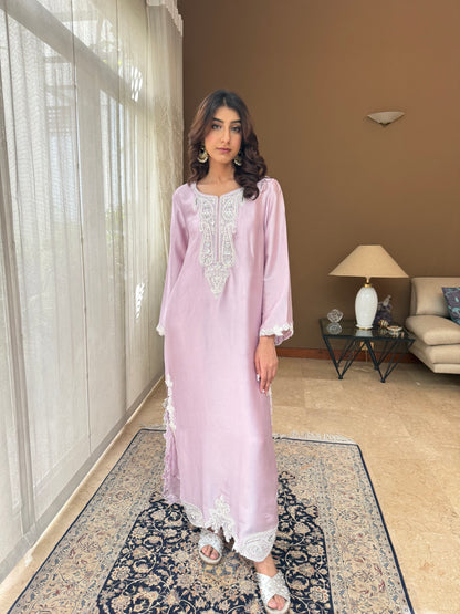 Woman in a light purple dress standing in a room with a beige wall and a patterned rug.